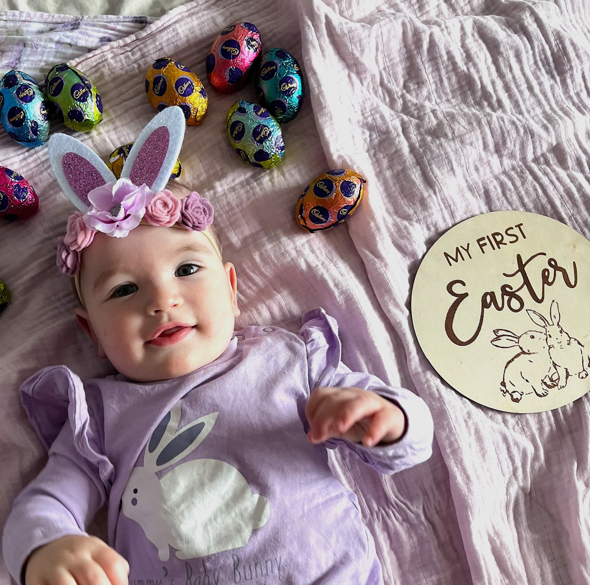 Baby wearing bunny ears and a 'My First Easter' sign with colourful Easter eggs on a pink blanket.