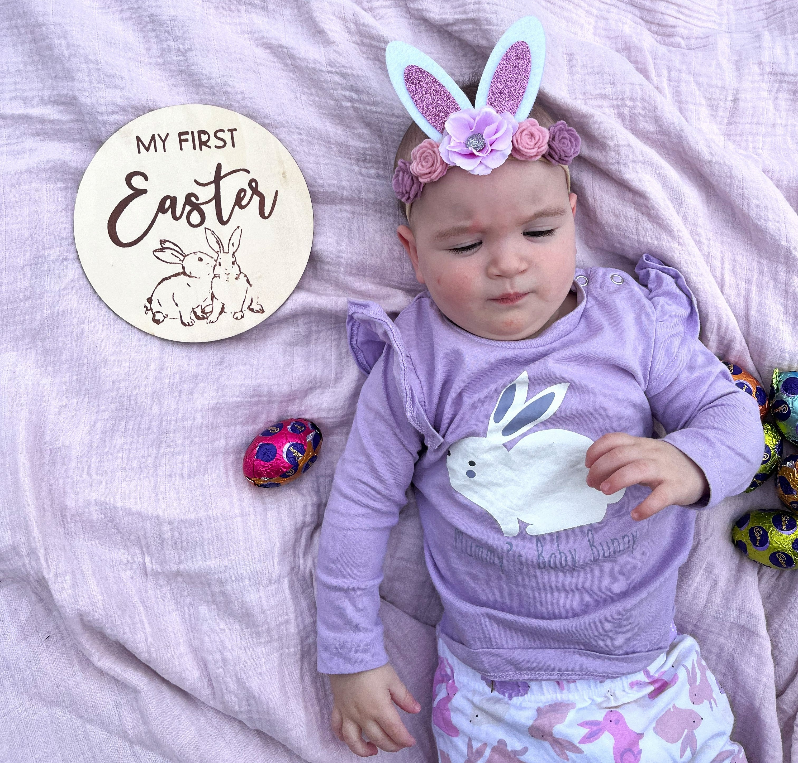 Baby wearing a bunny ears headband and a purple bunny top, surrounded by Easter eggs on a pink blanket