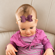 Baby wearing a purple headband with a bow, sitting on a beige couch.