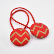 A pair of red fabric covered buttons with gold chevron patterns, attached to red elastic hair ties.